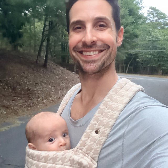 David smiling outdoors, carrying his baby son in a carrier